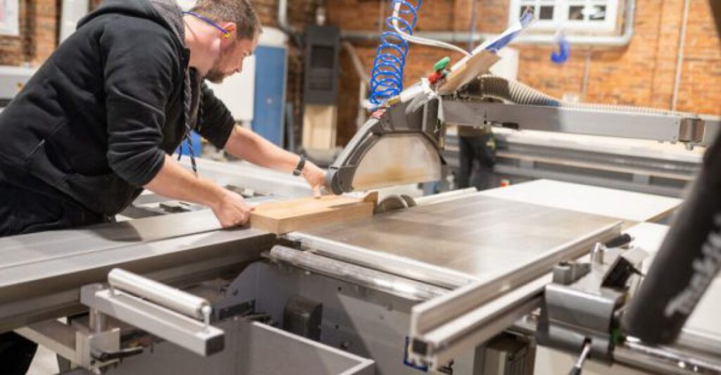 Ein Mann mit Bart arbeitet konzentriert an einer Kreissäge in einer Werkstatt, trägt schwarze Kleidung.