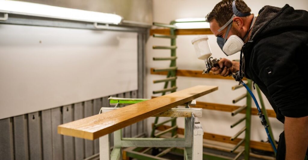 Ein Mann sprüht lackierte Holzlatte an, trägt Schutzmaske und schwarze Kleidung.