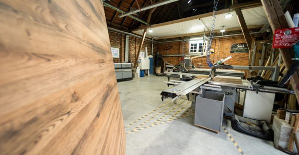 Große Tischlerei mit Holzbearbeitungsmaschinen, braune Holzwand links, Ziegelwände im Hintergrund.