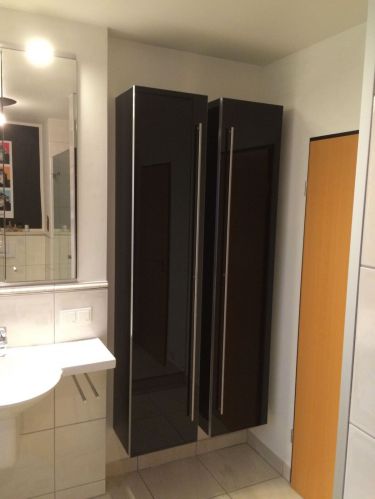 Schwarze Badezimmerschränke an weißer Wand, daneben ein Spiegel und ein weißes Waschbecken.