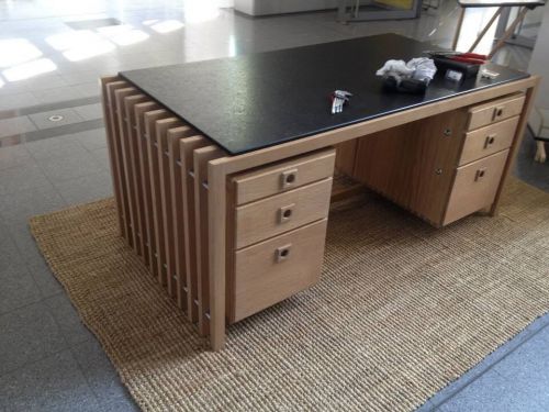 Moderner Holztisch auf Teppich mit Schubladen, schwarze Tischplatte, Bürogegenstände darauf.