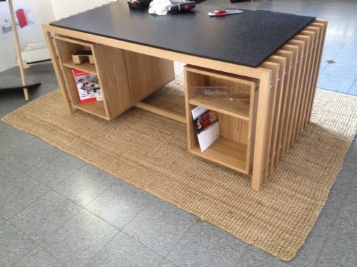 Moderner Holztisch mit Regalen, schwarze Oberfläche, auf geflochtener Matte, graue Bodenfliesen.