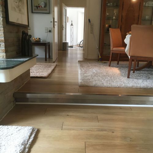 Wohnzimmer mit Holzboden, rote Stühle, Teppiche, Glastür, Besen, Staubsauger im Hintergrund.