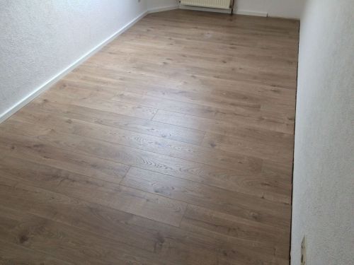 Leerer Raum mit hellem Holzfußboden, weißen Wänden und kleinem Heizkörper im Hintergrund.