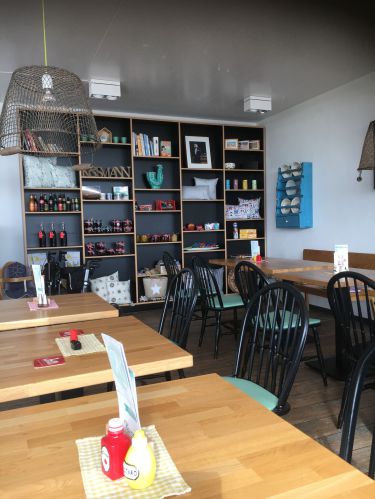 Gemütliches Café mit Holztischen, schwarzen Stühlen und einem Regal voller Bücher und Dekorationen.