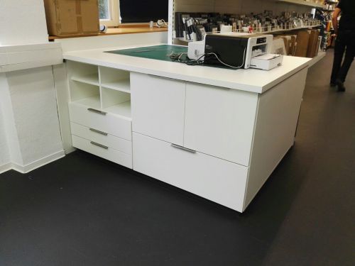 Weißer Schreibtisch mit Schubladen, Drucker darauf, in einem Büro mit dunklem Boden.