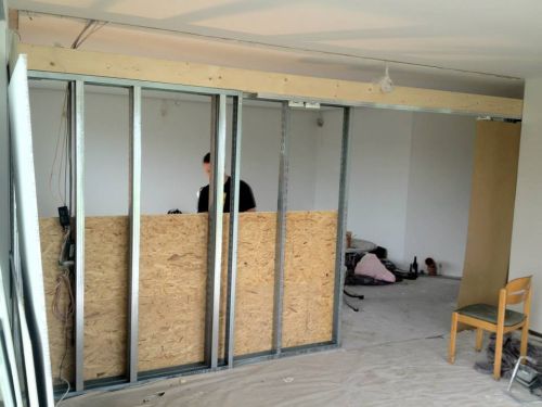 Eine Person arbeitet an einer teilweise errichteten Wand aus Metall und Holz in einem Raum.